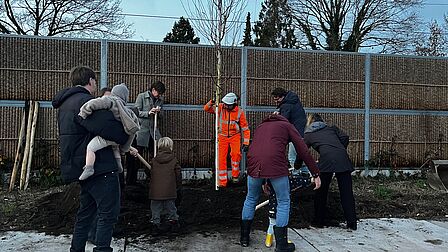 Mensen met schep graven gat voor boom en mensen kijken toe waarvan een kind en iemand met kind op arm