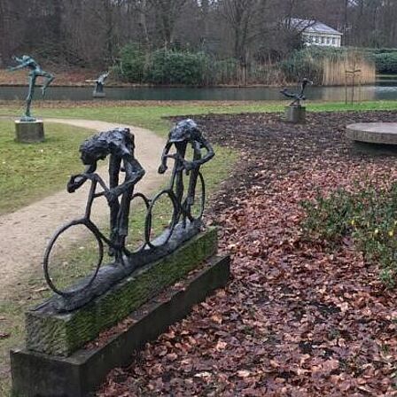 Inkijk Beeldenpark Jits Bakker op landgoed Beerschoten met een wandelpad langs gras en struiken, een vijver. Op de voorgrond een beeld van twee wielrenners, op de achtergrond nog 3 beelden.